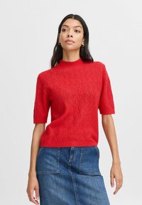 Roter Strickpullover mit hohem Kragen und kurzen Ärmeln. Der Stoff hat ein strukturiertes Wellenmuster. Getragen mit einem Jeansrock.