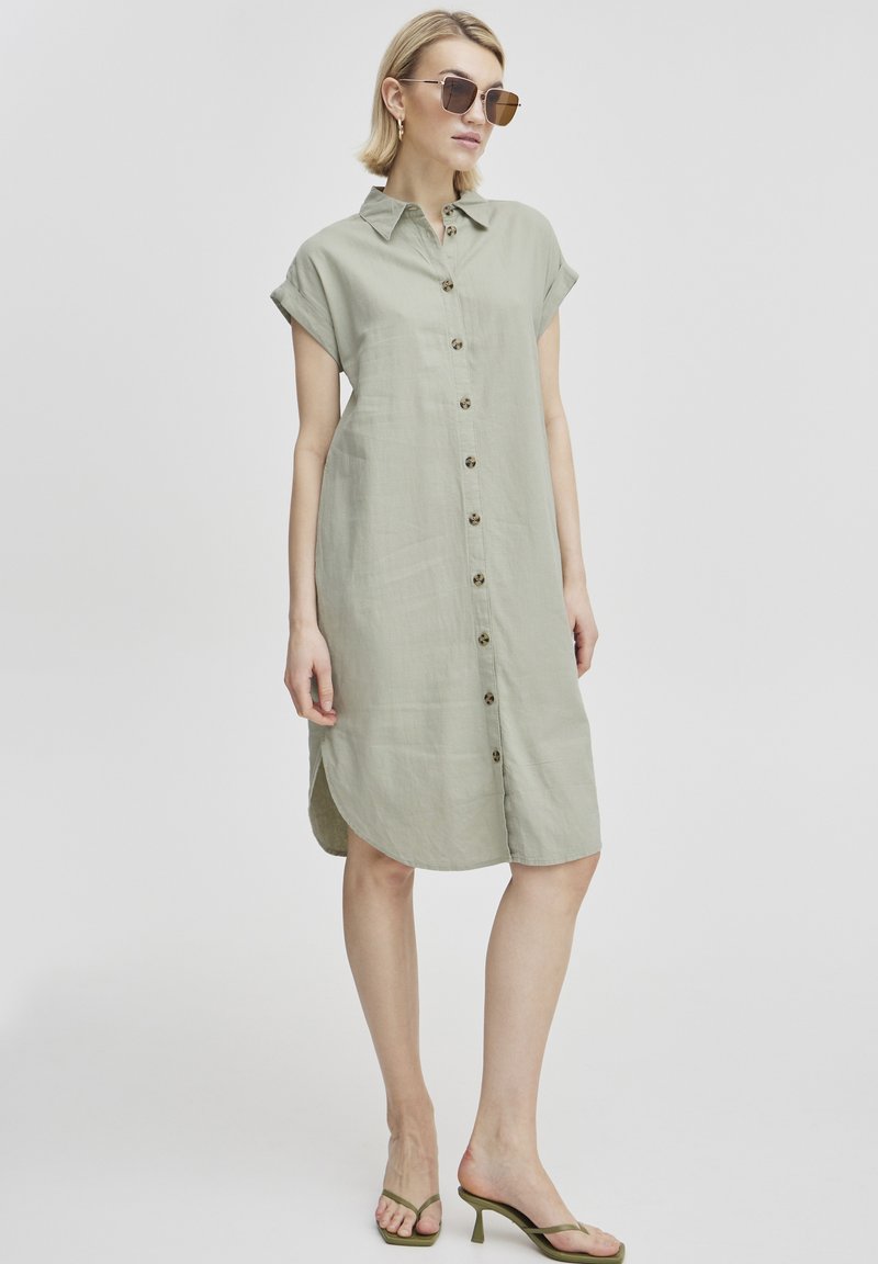Abito button-up verde oliva chiaro con colletto e maniche corte, caratterizzato da un taglio dritto, orlo arrotondato e dettagli con bottoni in legno.