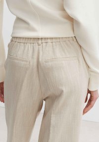 Beige strukturierte Hose mit elastischem Bund, ausgestattet mit zwei Gesäßtaschen mit flachen Nähten, kombiniert mit einem hellen Oberteil.