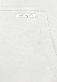 Sweat-shirt gris clair avec une poche avant et une étiquette argentée portant l'inscription "bazix republiq." Texture douce et design minimaliste.