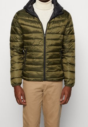 Mand iført olivengrøn quiltet jakke med sort for, hvid rullekravegenser og beige bukser, stående mod en ensfarvet baggrund.