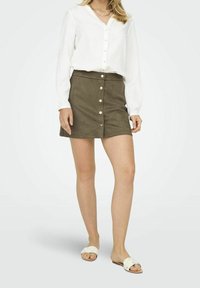 Falda de ante verde oliva con detalle de botones delanteros, combinada con una blusa blanca de manga larga. La modelo lleva sandalias planas blancas.