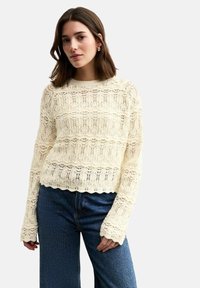 Pull en crème tricoté, présentant un motif texturé en openwork avec des bords festonnés, associé à un jean bleu taille haute à jambes larges.