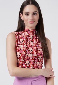 Top floral de cuello alto en rojo con flores rosa y negras. Diseño sin mangas, tejido suave, llevado con pantalones morados de talle alto.