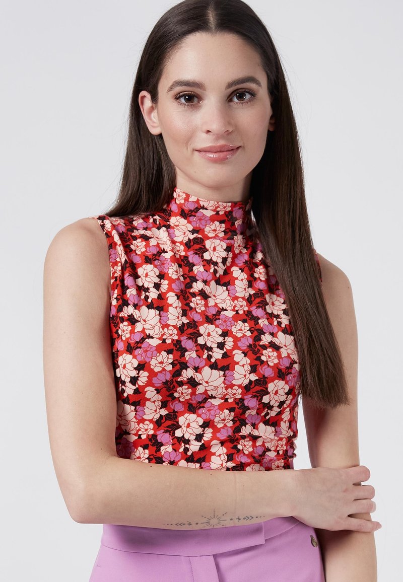 Top floral de cuello alto en rojo con flores rosa y negras. Diseño sin mangas, tejido suave, llevado con pantalones morados de talle alto.