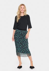 Haut noir à côtes avec des manches trois-quarts, associé à une longue jupe transparente ornée d'un motif floral vert et bleu sur fond noir.