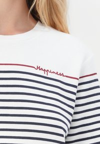 Witte sweatshirt met marineblauwe horizontale strepen. Bevat de tekst "Happiness" in het rood geborduurd op de borst. Zachte textuur en een ronde hals.