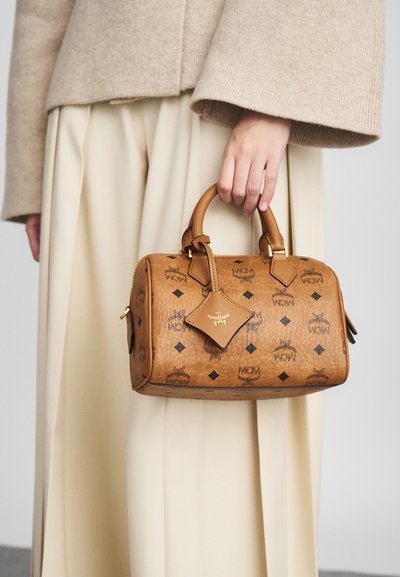 Hånd holder en medium brun læderhåndtaske med sorte logo prints, guldbeslag og rund håndtag, iført beige lange ærmer og cremefarvede bukser.