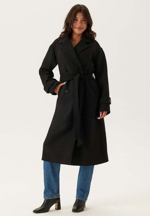 Sort uldtrenchcoat med bæltestrik, dobbeltbrystede knapper og brede revers. Båret over blå jeans og sorte støvler med hæl.