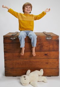 Maglione giallo con giromanica decorato, jeans blu e piedi nudi. Bambino seduto su una cassa di legno con un orsetto di peluche bianco sul pavimento.