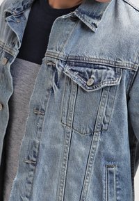 Hellblaue Jeansjacke mit verblasster Textur, zwei Brusttaschen, Knopfleiste vorne und kontrastierender grauer Innenlage.