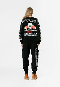 Persona con cabello largo viste un sudadera negra con un cartoon y texto blanco, pantalones cargo negros con parches y botas color beige, de pie de espaldas.