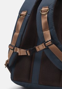 Sac à dos en tissu bleu foncé, avec des panneaux en maille marron et des accessoires en bronze. Dispose de bretelles rembourrées et d'une boucle ajustable pour le soutien.