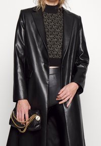 Cappotto di pelle nera su un top in maglia nera testurizzata con accenti dorati. Tiene in mano una piccola borsa nera con una tracolla dorata e logo.