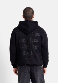 Felpa nera con cappuccio e una grande scritta testurizzata sul retro. Presenta un cappuccio con lacci e polsini a costine. Design semplice, vestibilità casual.