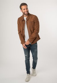 Bruine suede jas met een knoopsluiting aan de voorkant, twee borstzakken en een gladde textuur, gepaard met versleten blauwe jeans en witte sneakers.