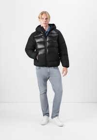 Doudoune noire avec capuche, présentant une finition brillante et un matelassage horizontal, assortie à un jean bleu clair et des baskets blanches.