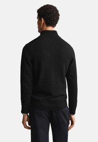 Hombre visto desde atrás que lleva un suéter de punto acanalado negro con cuello alto y pantalones oscuros sobre un fondo blanco liso.