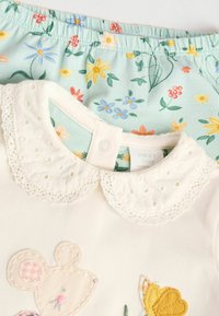 Weißes Babyoberteil mit Spitzenkragen, das bestickte Maus- und Schmetterlingsmuster aufweist; kombiniert mit kurzen Hosen im grünen Blumenmuster.