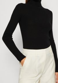 Maglione nero a coste con colletto a imbuto e maniche lunghe, design aderente, indossato con pantaloni a vita alta di colore avorio. Tessuto liscio e materiale elasticizzato.