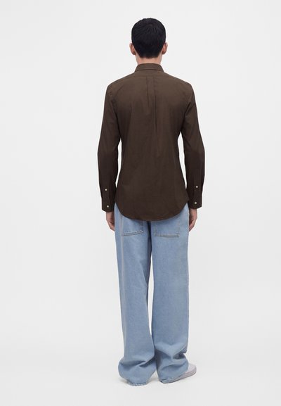 Homme portant une chemise marron foncé à manches longues et un jean large bleu clair, debout de dos sur un fond blanc.