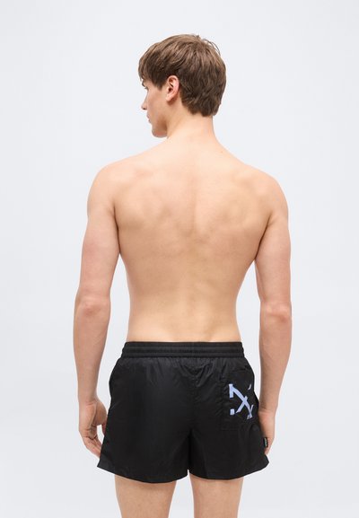 Jeune homme vu de dos portant un short de bain noir avec un motif de flèche blanche sur la poche arrière, sur un fond uni.