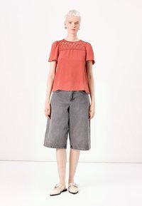 Top corto color ruggine con un pannello in pizzo testurizzato. Abbinato a culotte in denim grigio e scarpe slip-on beige. Vestibilità semplice e rilassata.