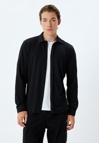 Zwarte langarmshirt met een button-down kraag, textuurstof en sluiting met knopen aan de voorkant. Draag het over een wit T-shirt.