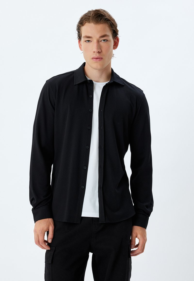 Zwarte langarmshirt met een button-down kraag, textuurstof en sluiting met knopen aan de voorkant. Draag het over een wit T-shirt.