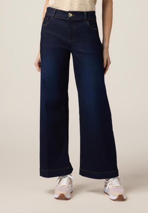 Jean large bleu foncé en denim, avec une fermeture à bouton à l'avant, des passants de ceinture et des détails de couture subtils. Associé à des baskets claires.