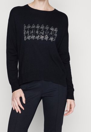 Person iført en sort sweater med et sølvfarvet hanefjedmønster på brystet og sorte bukser, stående mod en ensfarvet lys baggrund.