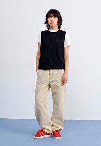Svart stickad väst över vit t-shirt, matchad med lösa beige byxor och röda sneakers. Bakgrunden är helt vit med ett blått golv.
