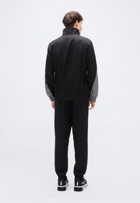 Μαύρη φόρμα με ψηλό γιακά και γκρι λεπτομέρειες στα μανίκια, με παντελόνια που στενεύουν προς τα κάτω και ριμπ μανσέτες. Απαλό, ελαφρύ ύφασμα.