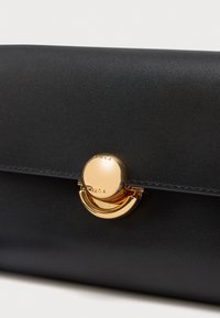 Fekete bőr pénztárca, kerek, fényes arany csattal, amelyre a "Furla" márkanevet vésték, egy egyszerű, világos háttéren.