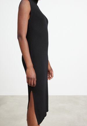 Robe noire sans manches en maille côtelée avec fente latérale portée par une personne debout devant un fond uni.