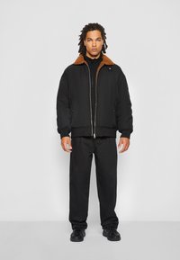 Veste noire matelassée avec un col en fausse fourrure marron, fermeture éclair, poignets côtelés, portée sur un col roulé noir, assortie à un pantalon noir et des baskets.