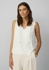 Weiße ärmellose Bluse mit V-Ausschnitt, verziert mit filigraner Blumenstickerei und einer glatten Textur. Kombiniert mit cremefarbenen Hosen.
