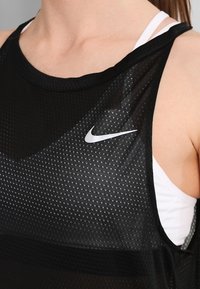 Primer plano de una persona que lleva una camiseta sin mangas negra de malla de Nike sobre un sujetador deportivo blanco, mostrando el logo de Nike en el pecho.