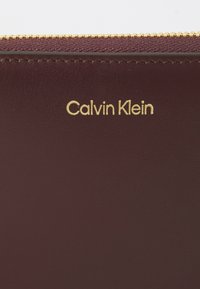 Portofel din piele burgundy cu logo-ul aurie "Calvin Klein", având o finisare texturată și o fermoar aurie pe marginea de sus.