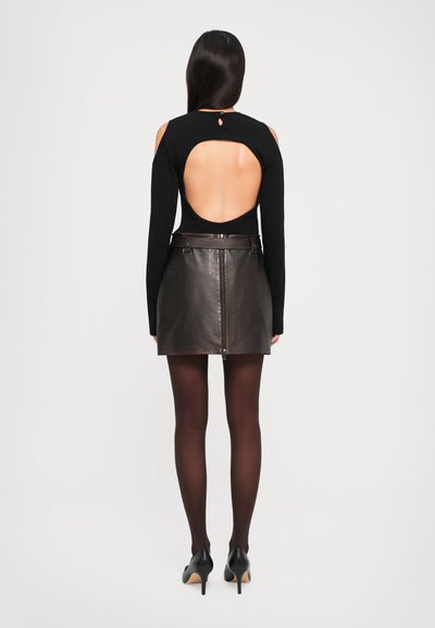 Woman standing facing away, wearing black long-sleeve top with open back, dark brown leather skirt with zipper, black tights, and black heels.