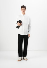 Ung mann står mot hvit vegg, kledd i hvit skjorte med knapper, svarte bukser, hvite joggesko, og holder en svart veske under den ene armen.