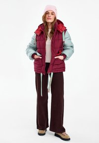 Nő áll, rózsaszín sapkában, bordó és világoskék steppelt dzsekiben, bézs pulóverben, barna bőszárú nadrágban és barna túrabakancsban.