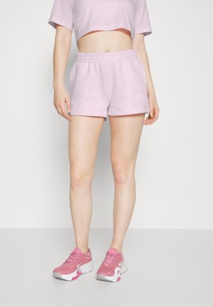 Personne portant un short violet clair et un haut court avec des chaussures de sport roses et blanches, debout sur un fond uni.