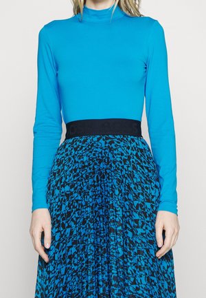 Femme portant un haut à col roulé bleu vif ajusté à manches longues et une jupe plissée bleu et noir à motif, avec une large ceinture noire.