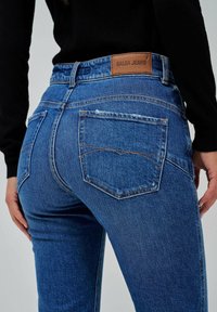 Jean oscuro de mezclilla con un diseño ajustado, que cuenta con dos bolsillos traseros con costuras en contraste y una etiqueta de cuero marrón en la cintura.