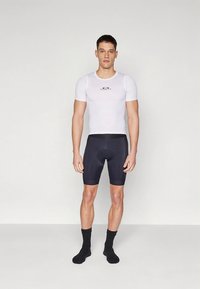 Valkoinen lyhythihainen urheilupaita, jossa on logo, yhdistettynä mustiin pyöräilyshortseihin. Asu on vartalonmyötäinen, valmistettu joustavasta, hengittävästä kankaasta.