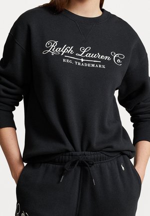 Person iført sort sweatshirt og matchende joggingbukser med hvid "Ralph Lauren Co" tekst og logo, hænderne i lommerne.