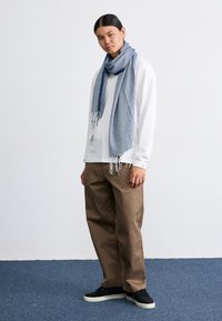 Blue scarf with a textured pattern, fringed edges, draped over a white long-sleeve shirt, paired with brown pants and black shoes.