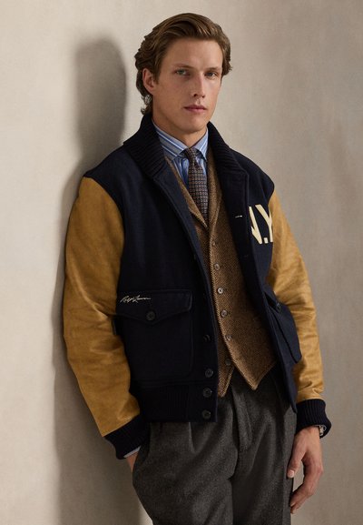 Ναυτικό μαλλινό σακάκι με μπεζ σουέντ μανίκια, ραβδωτό γιακά και κουμπωτή μπροστινή πλευρά. Περιλαμβάνει τσέπη στο στήθος και λεπτομέρεια λογότυπου, φορεμένο πάνω από γκρι καρό γιλέκο.