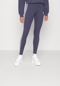 Námořnické modré legíny s bílými bočními pruhy, vyrobené z elastické tkaniny. Spárovány s bílými teniskami pro sportovní vzhled.